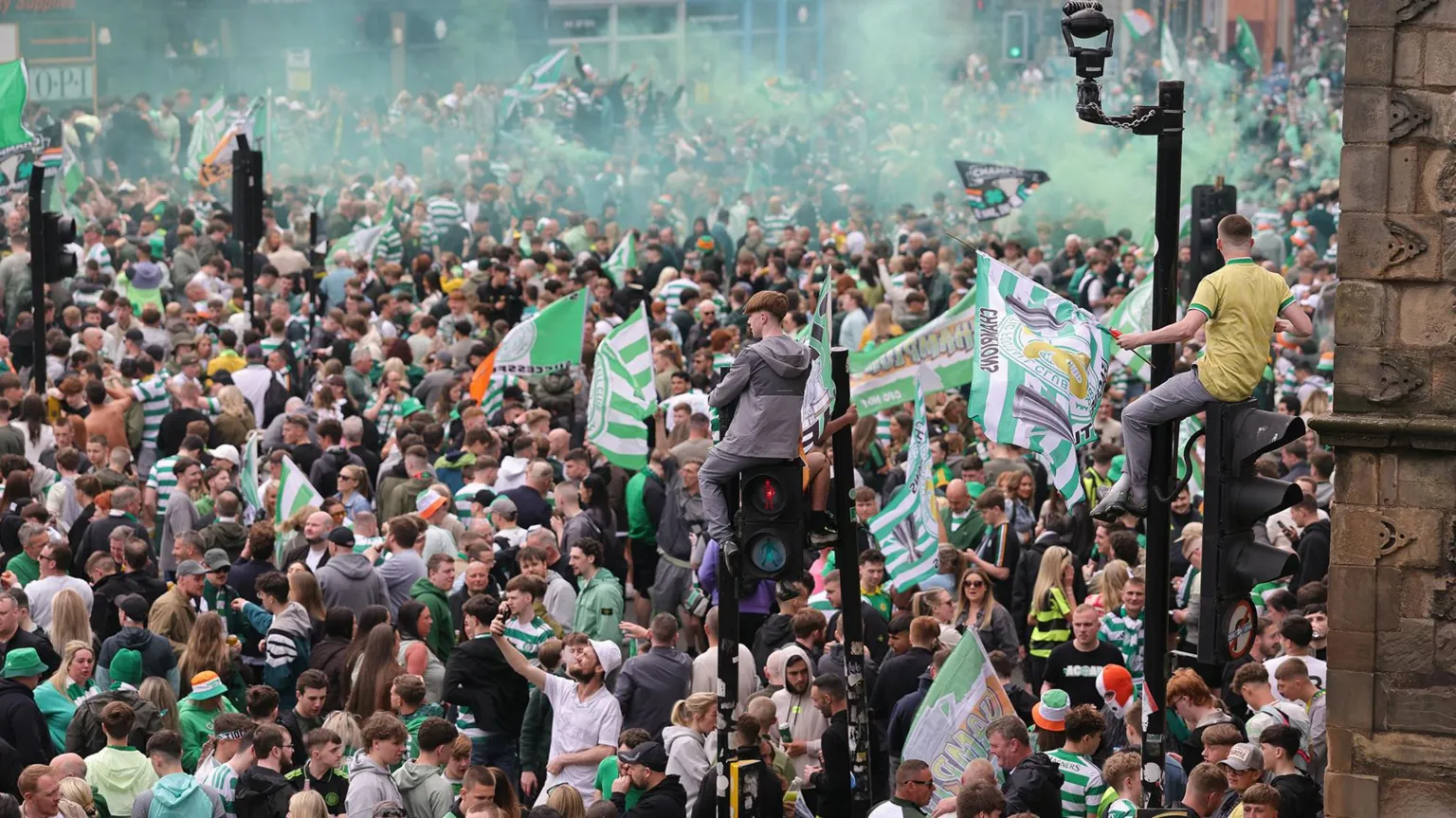  A large crowd of Celtic supporters gathers in a Glasgow street, with green smoke in the air and flags being waved, while some people sit on traffic lights.