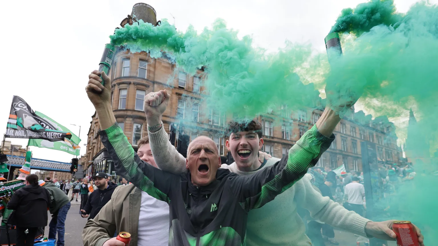 Three Celtic fans cheer for the camera while celebrating winning the league title at the weekend. One fan, a bald man in a track suit, has two smoke cannisters in his hand, which are letting off green smoke.