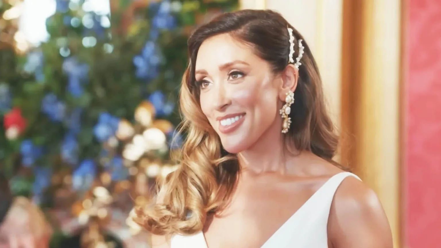 Married at First Sight UK, CPL Productions Shona stands in an elegant setting, wearing a white sleeveless wedding dress and ornate dangling earrings. She is captured side-on and smiling at her groom, who is off-camera. Her wavy light‑brown hair is styled with decorative pins on one side. Behind her, there is a blur of colourful flowers — blues, whites, golds and reds.