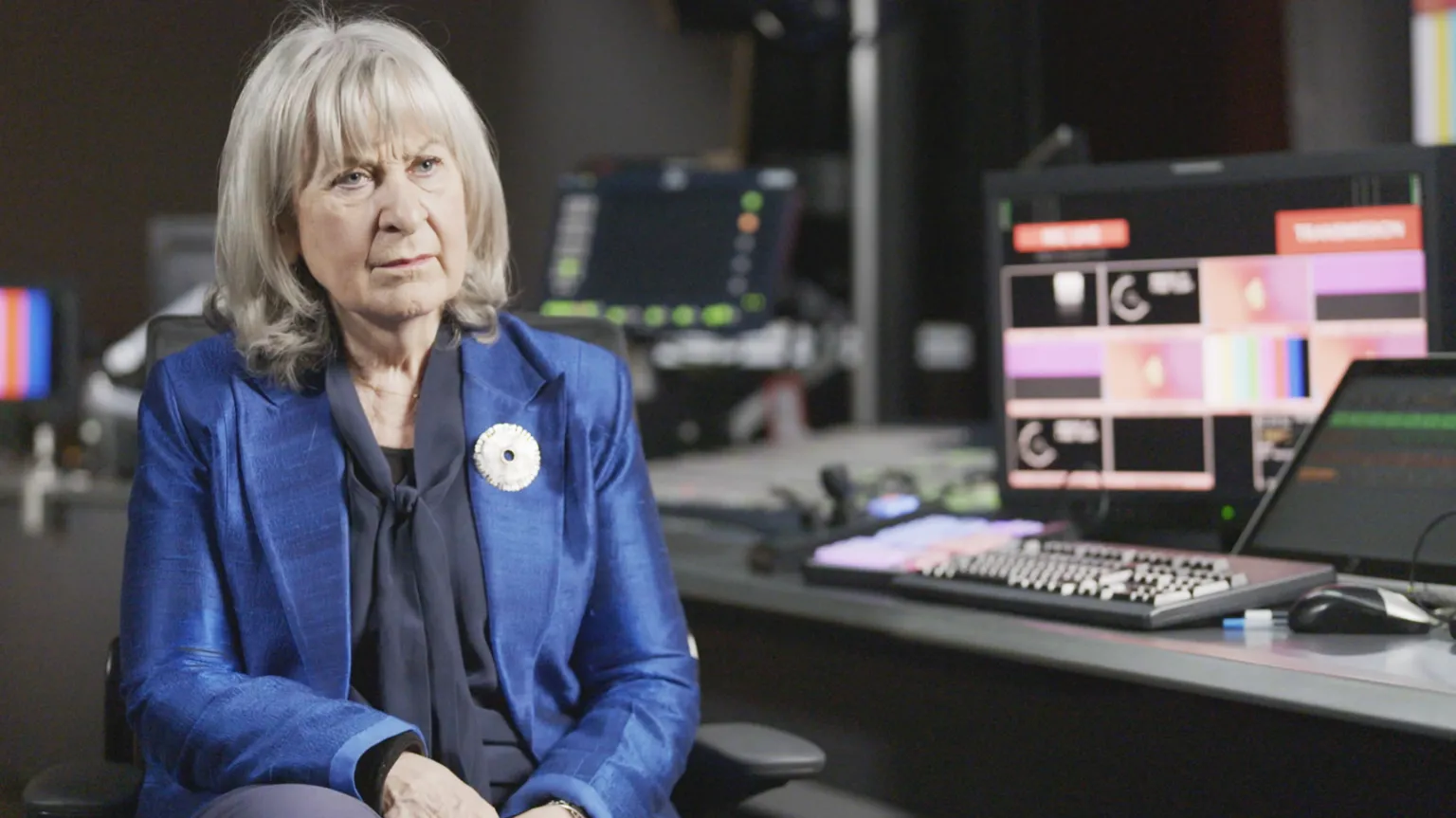 Baroness Helena Kennedy sits in a broadcast studio, wearing a bright blue blazer with a dark top and a white brooch. She has a pensive expression and behind her, several monitors glow with colourful test patterns and control screens, surrounded by keyboards and technical equipment.