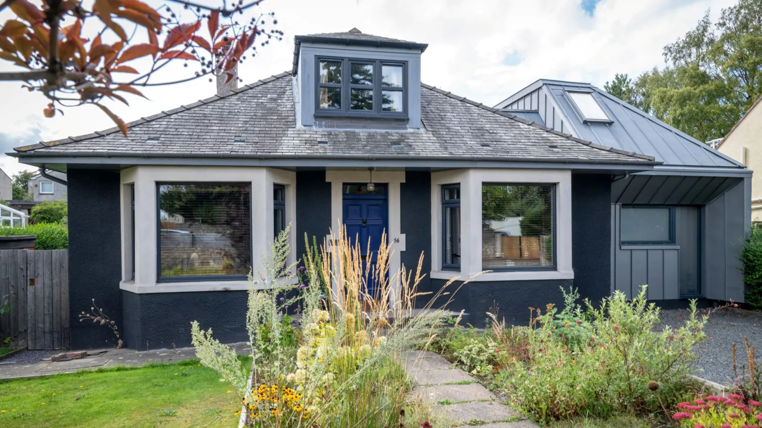 IWC Media An image of the front of a modernised 1960s bungalow. It is painted navy blue and has a modern side extension in grey materials. Rows of wildflower line the path to the front door which is a rich royal blue.