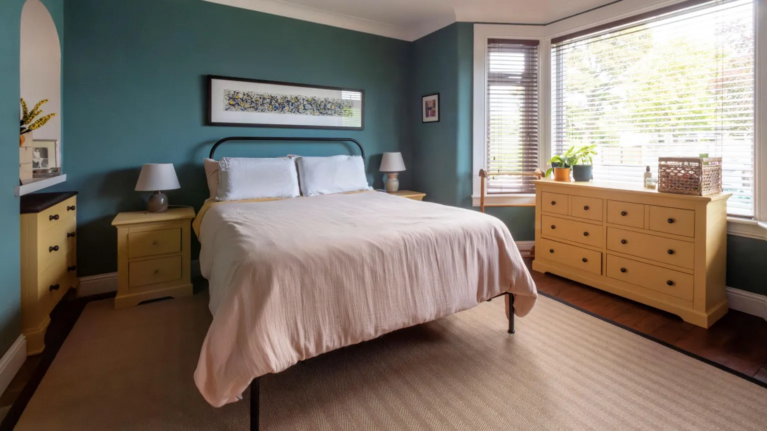 IWC Media A calm, neatly arranged bedroom features deep teal walls, a central bed with neutral bedding, and matching light wood bedside tables and drawers. Large bay windows with blinds let in natural light, complementing the warm tones of the wooden furniture and soft-textured rug.