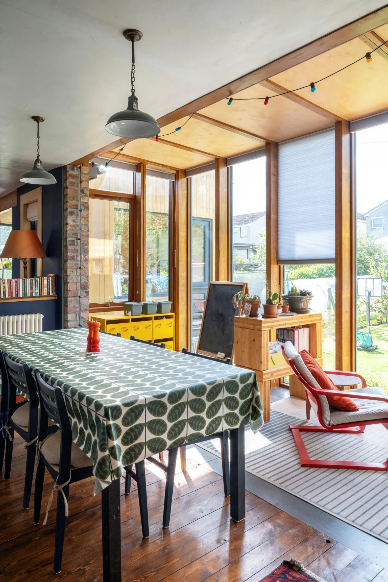 IWC Media A bright dining area sits within a sunlit extension, featuring large wooden-framed windows that look out onto a green garden. A rectangular table covered with a patterned cloth is surrounded by dark chairs, with pendant lights and colorful string lights hanging above. A mix of natural wood, exposed brick, houseplants, and shelving adds warmth and a relaxed, lived-in feel to the space.