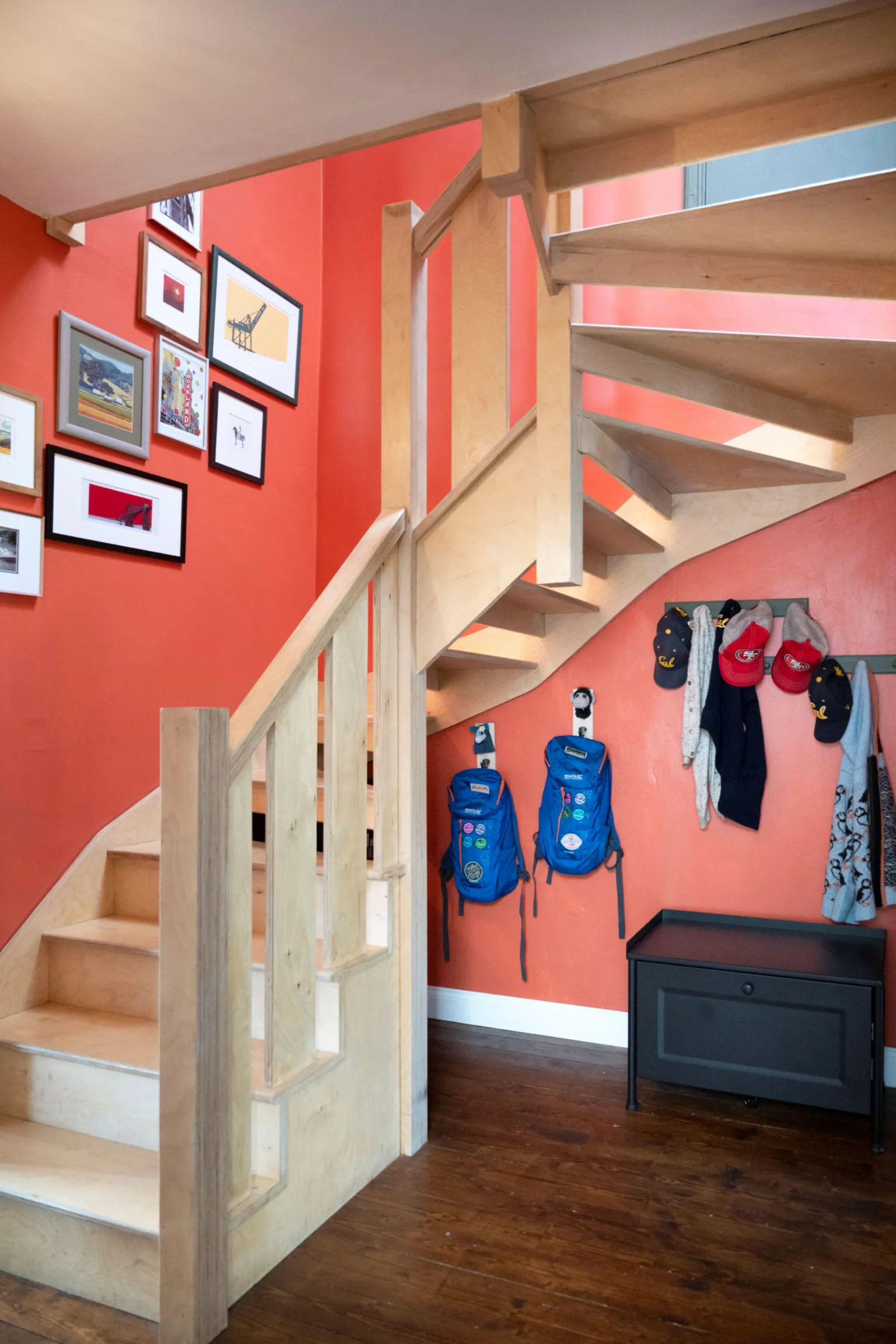 IWC Media A wooden staircase with open risers climbs against bold orange walls, creating a striking, modern interior focal point. Framed artwork decorates the wall, while hooks below hold jackets and backpacks above a small storage bench on polished wooden flooring.