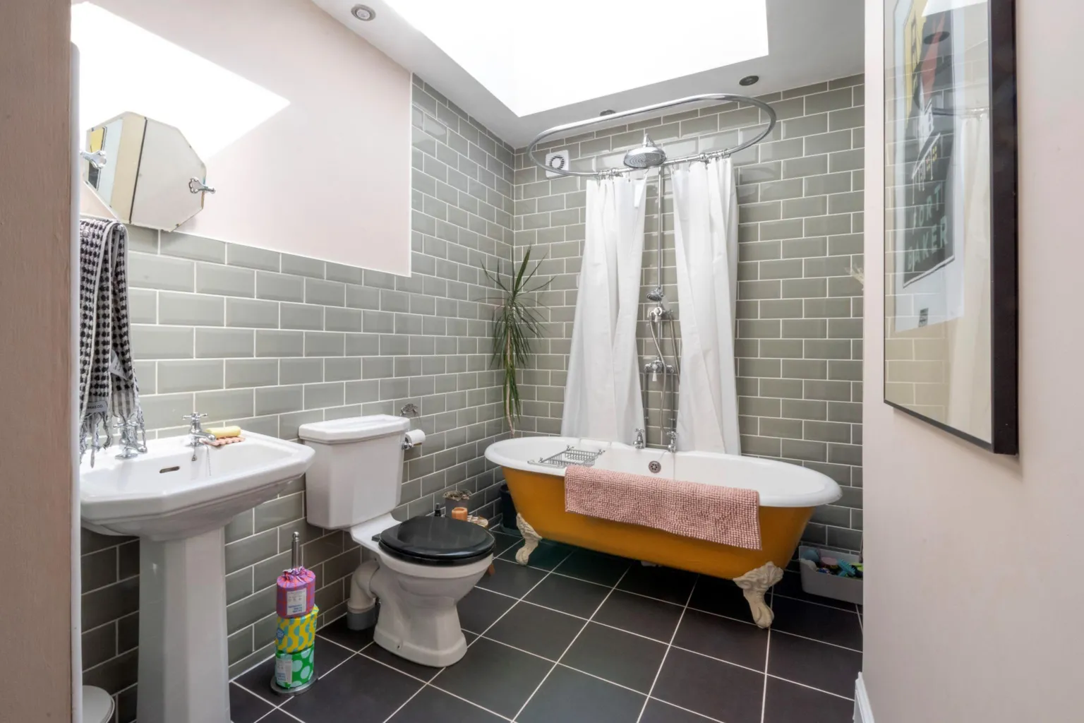 IWC Media A bright, modern bathroom features pale green subway tiles, a skylight, and a freestanding bathtub with a curved shower rail and white curtain. A white pedestal sink, toilet, and simple accessories sit against dark floor tiles, while soft daylight fills the compact space.