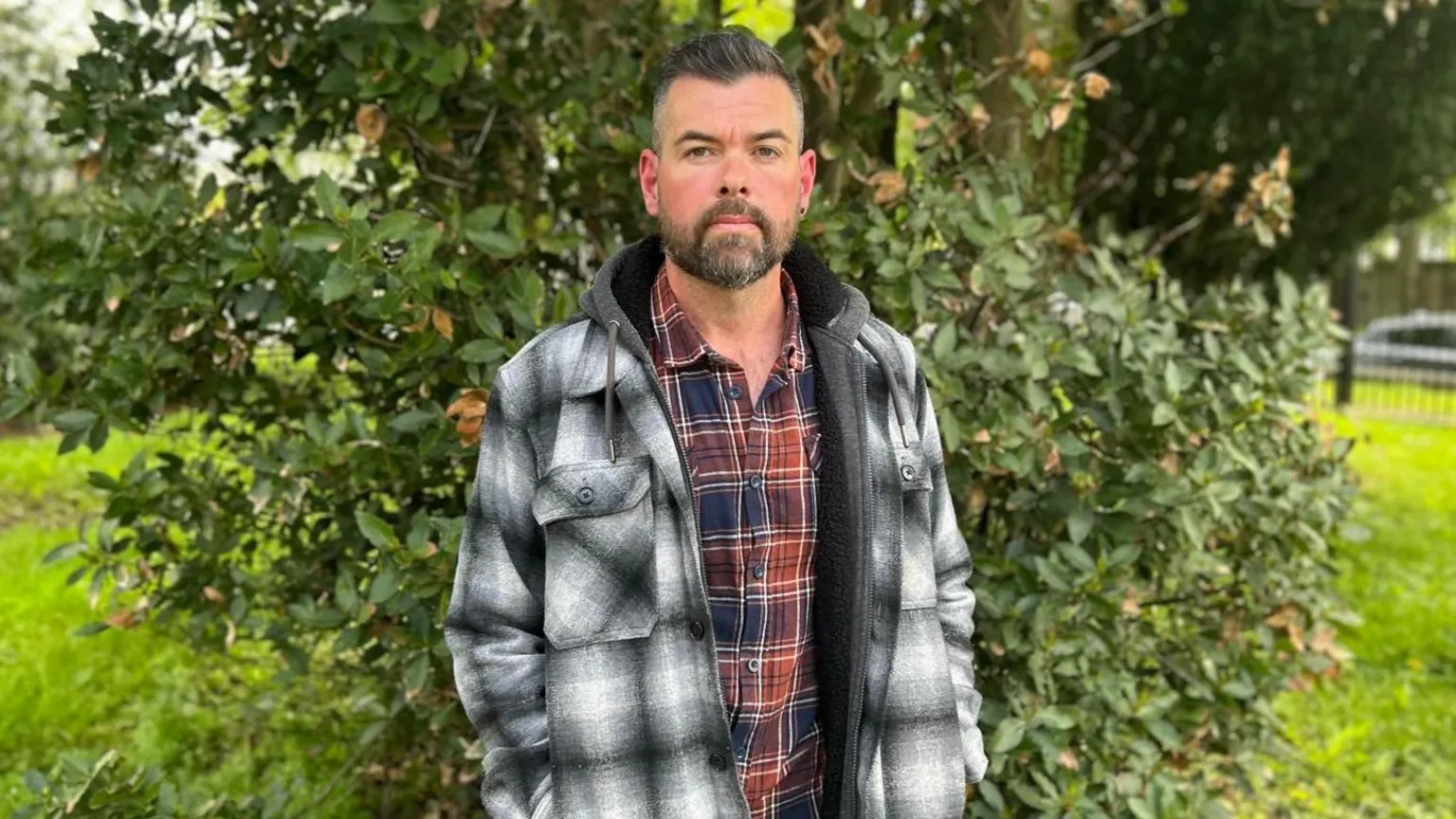 A man wearing a black and white checked jacket and red and blue checked shirt stands in front of shrubbery. He has a short beard, dark in colourand short dark hair. He is looking directly at the photographer.