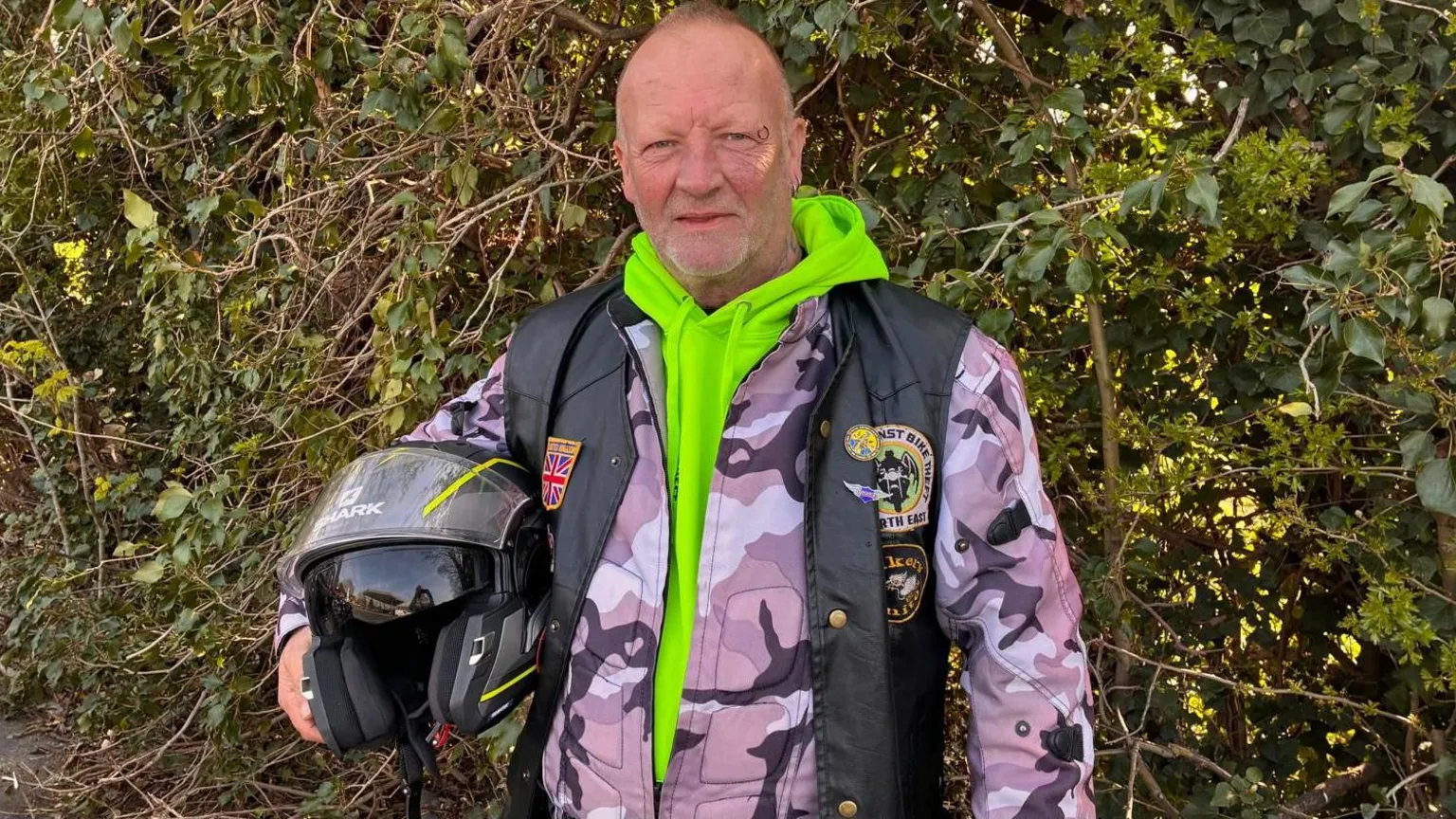 A man stands in front of a green hedge with a motorcycle helmet under his arm. He looks directly into the camera with a serious expression. He is wearing a black biking jacket over a combat-style coat and bright green hoody.