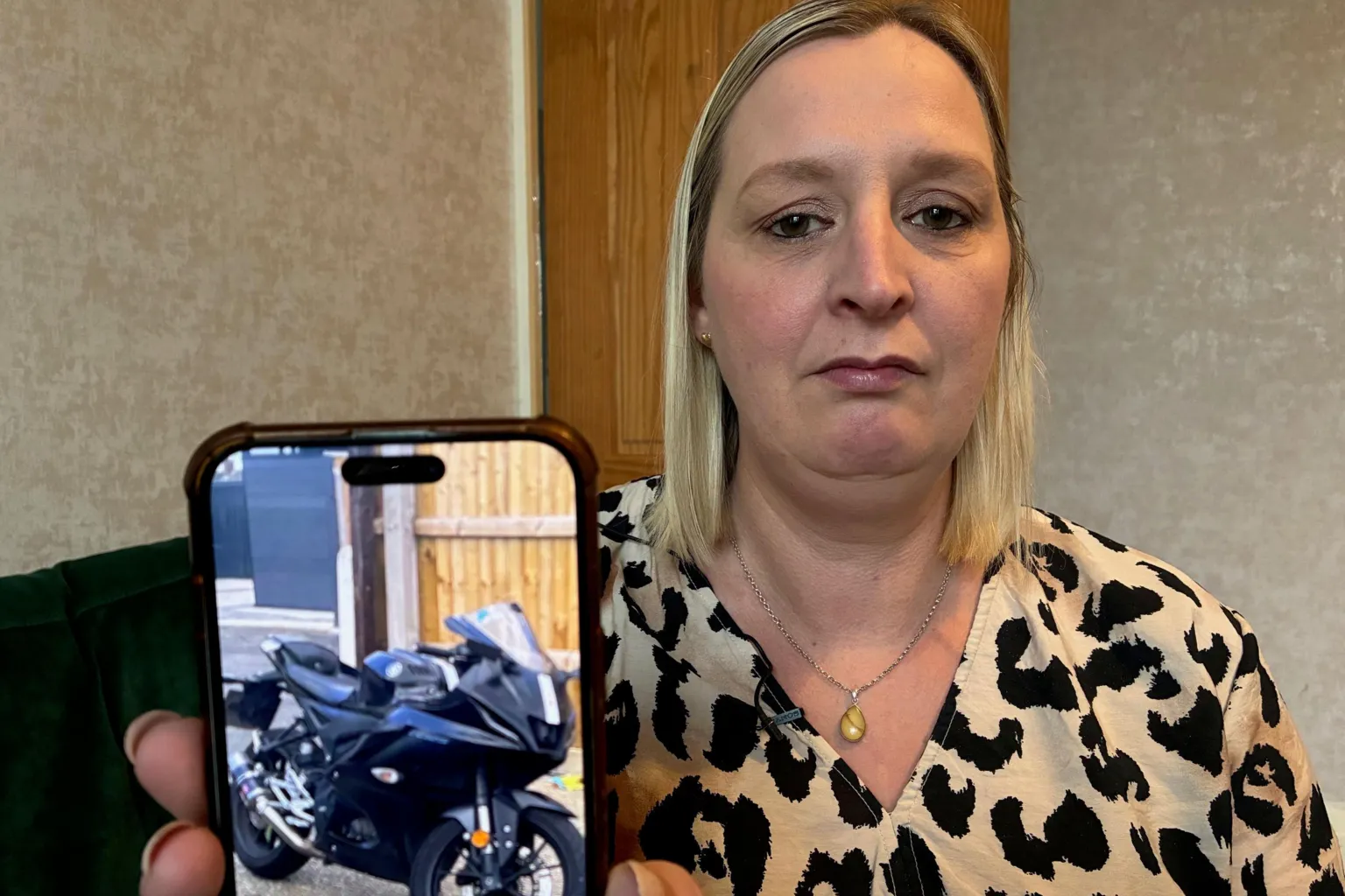 A woman with shoulder-length blond hair sits in a living room while holding her phone up to the camera. The phone screen shows a picture of a black motorbike. She wears a printed black and cream top and a silver pendant and has a serious expression on her face.