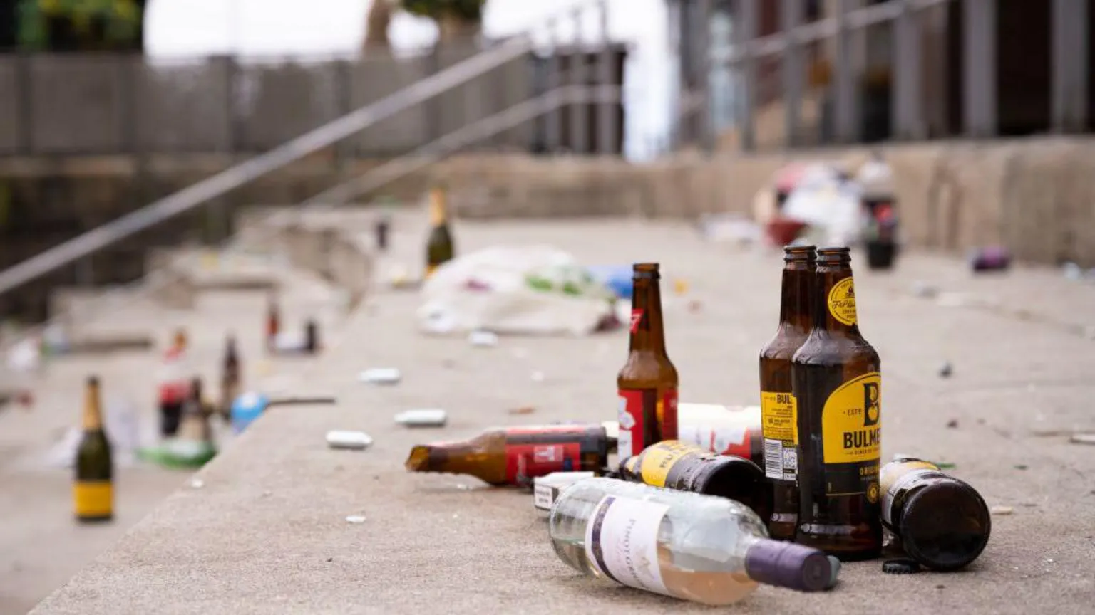A number of glass bottles can be seen scattered on a set of steps. There is wine bottle in the foreground alongside a few cider bottles. Just behind them, a smashed Budweiser beer bottle can be seen.