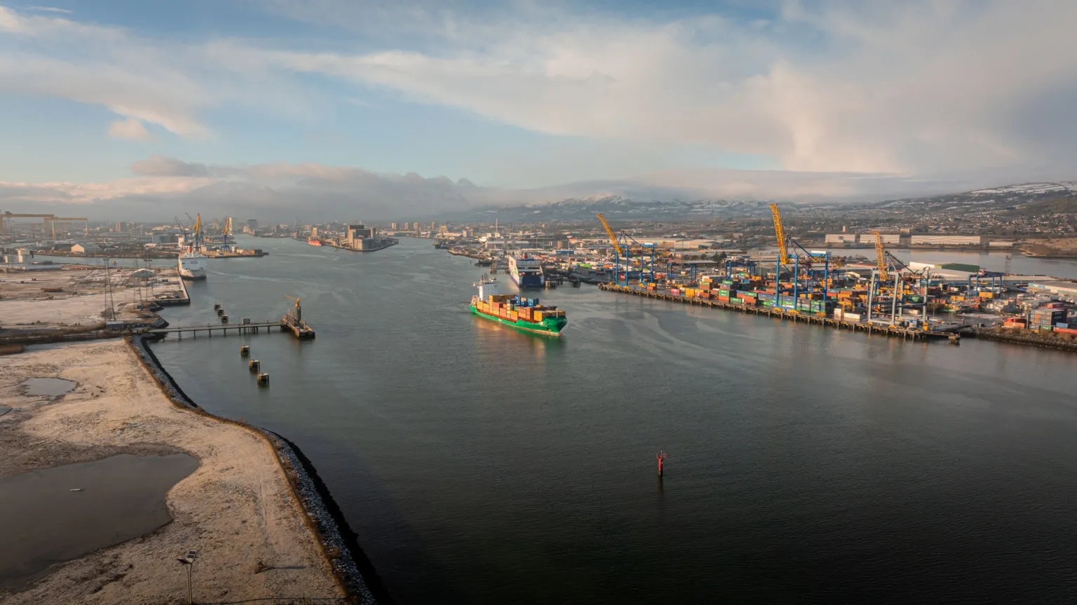 Belfast Harbour A ship pictured sailing with Belfast port in the background. The ship is green carrying yellow shipping containers.