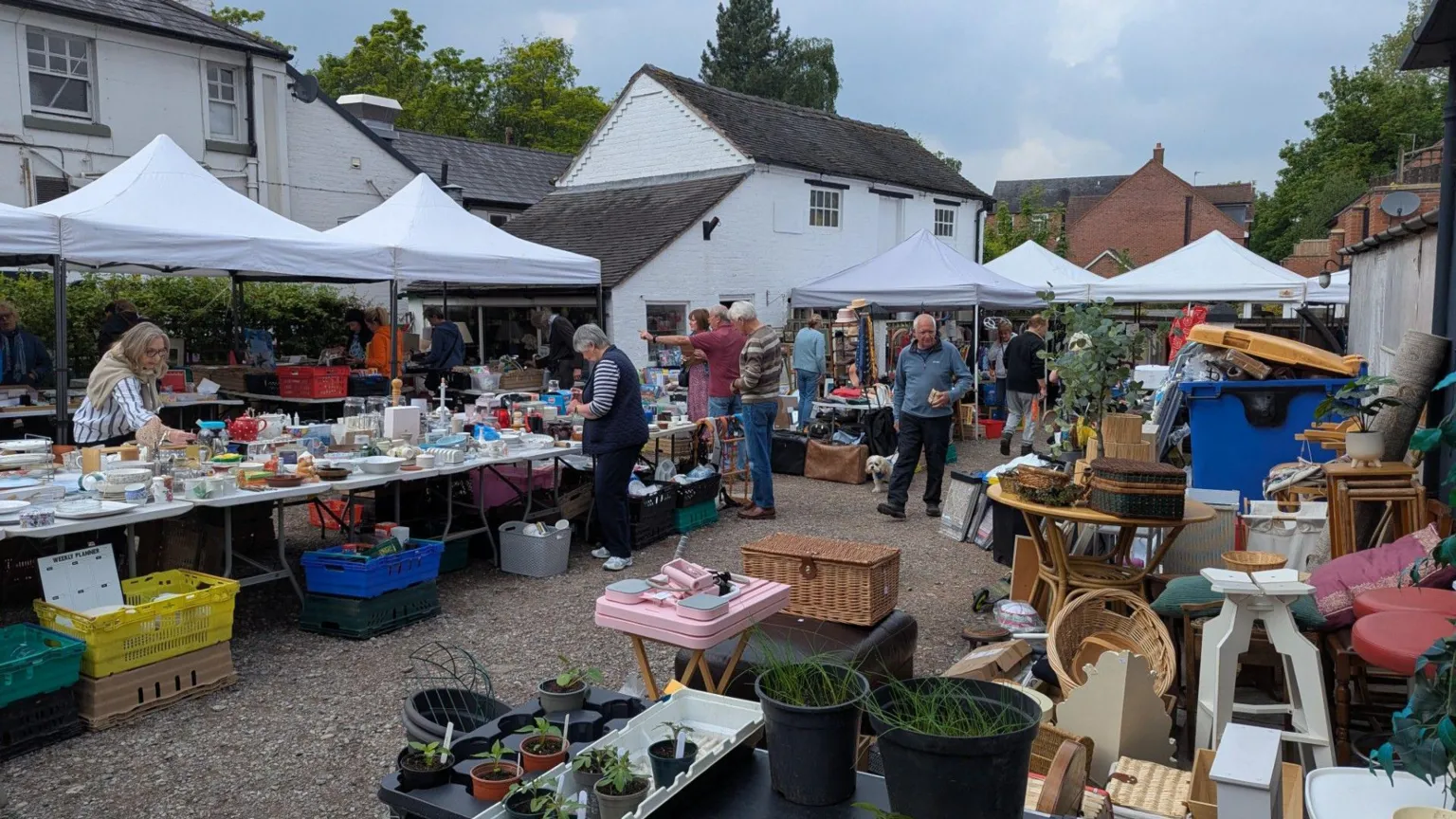 A number of covered stalls are laid out on ground outside a two storey white building.There are people browsing the goods, which are also laid out on the ground to the bottom right of the image