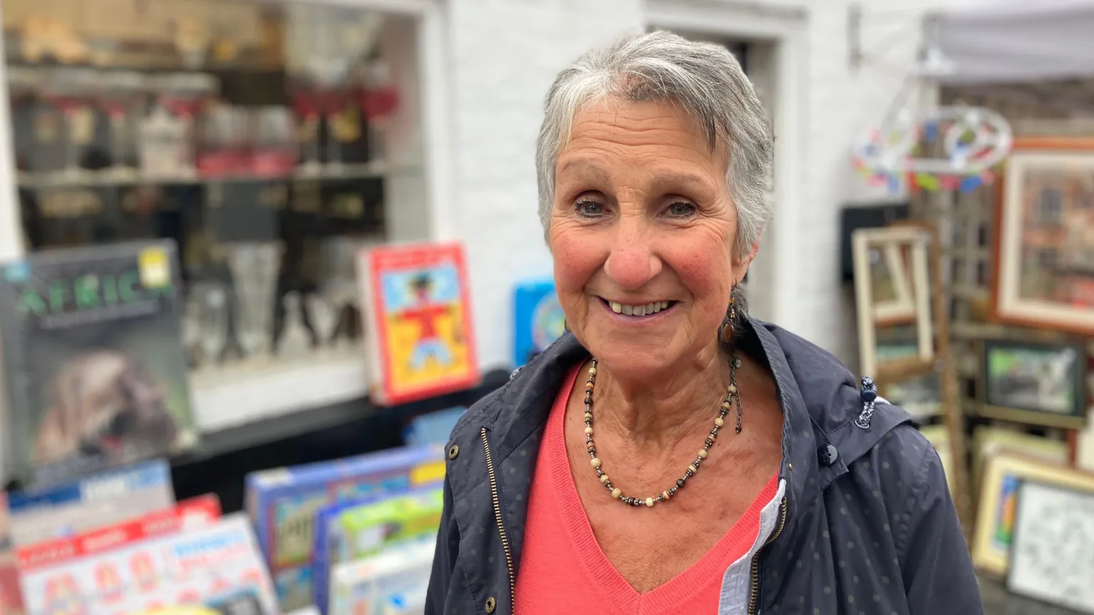A woman with grey hair, wearing a dark blue jacket over a pink top and necklace. She is smiling, and there are games and pictures displayed behind her