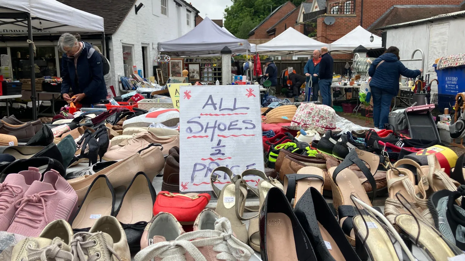 A close-up of pairs of shoes on a table with a sign saying all shoes half price. There are people browsing goods to either side of the table