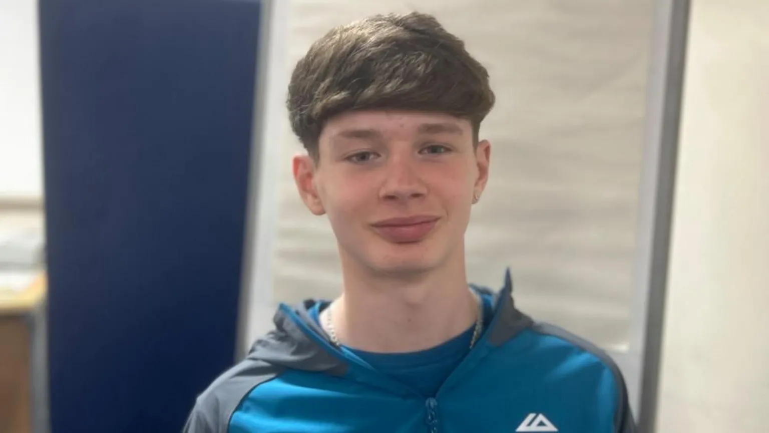Harrison is wearing a blue and grey jacket. He has longish brown hair and is smiling at the camera.