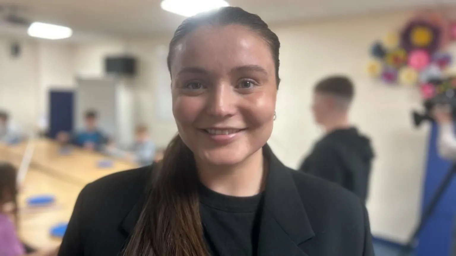 Zara is wearing a black suit jacket and black t-shirt. She has long brown hair tied back in a pony tail style. She is smiling at the camera.