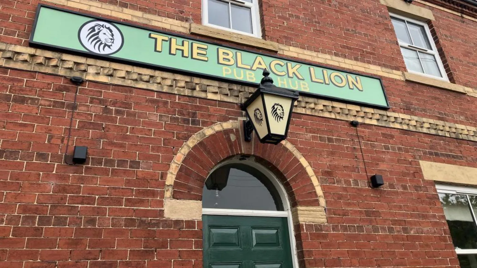 BBC / Fiona Callow A brick building facade features a green sign reading “The Black Lion Pub & Hub” with a lion logo. Below the sign is an arched doorway with a dark green door and a decorative lantern mounted above it. The structure includes textured brickwork patterns and windows on the upper floor, giving a traditional pub appearance.