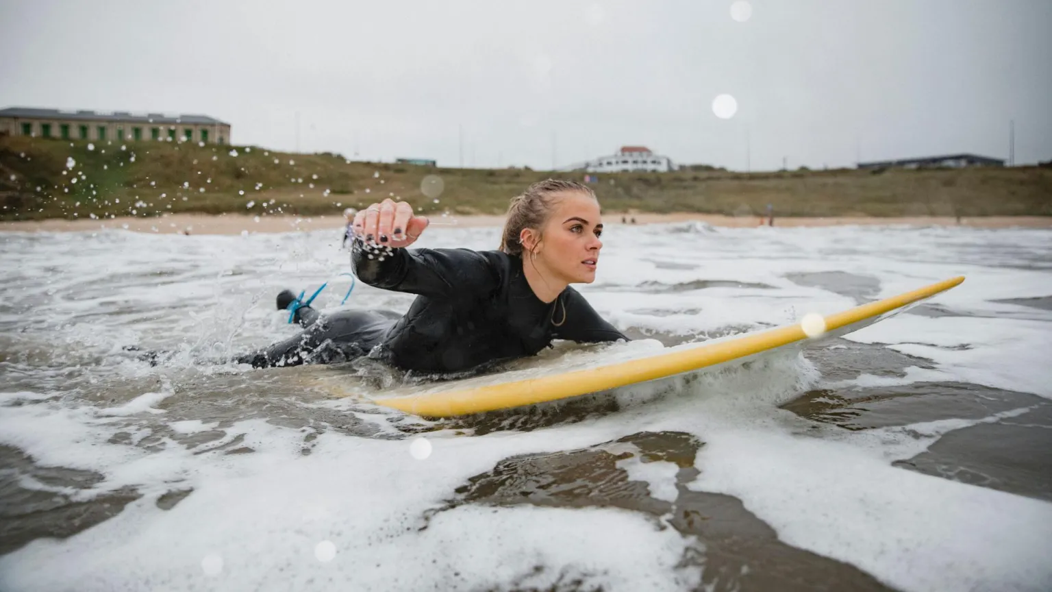  A young woman with long blonde hair lies on a yellow surfboard and paddles out on the sea. Behind her is the beach and a cliff atop of which sits several small buildings. The picture is taken at Tynemouth.