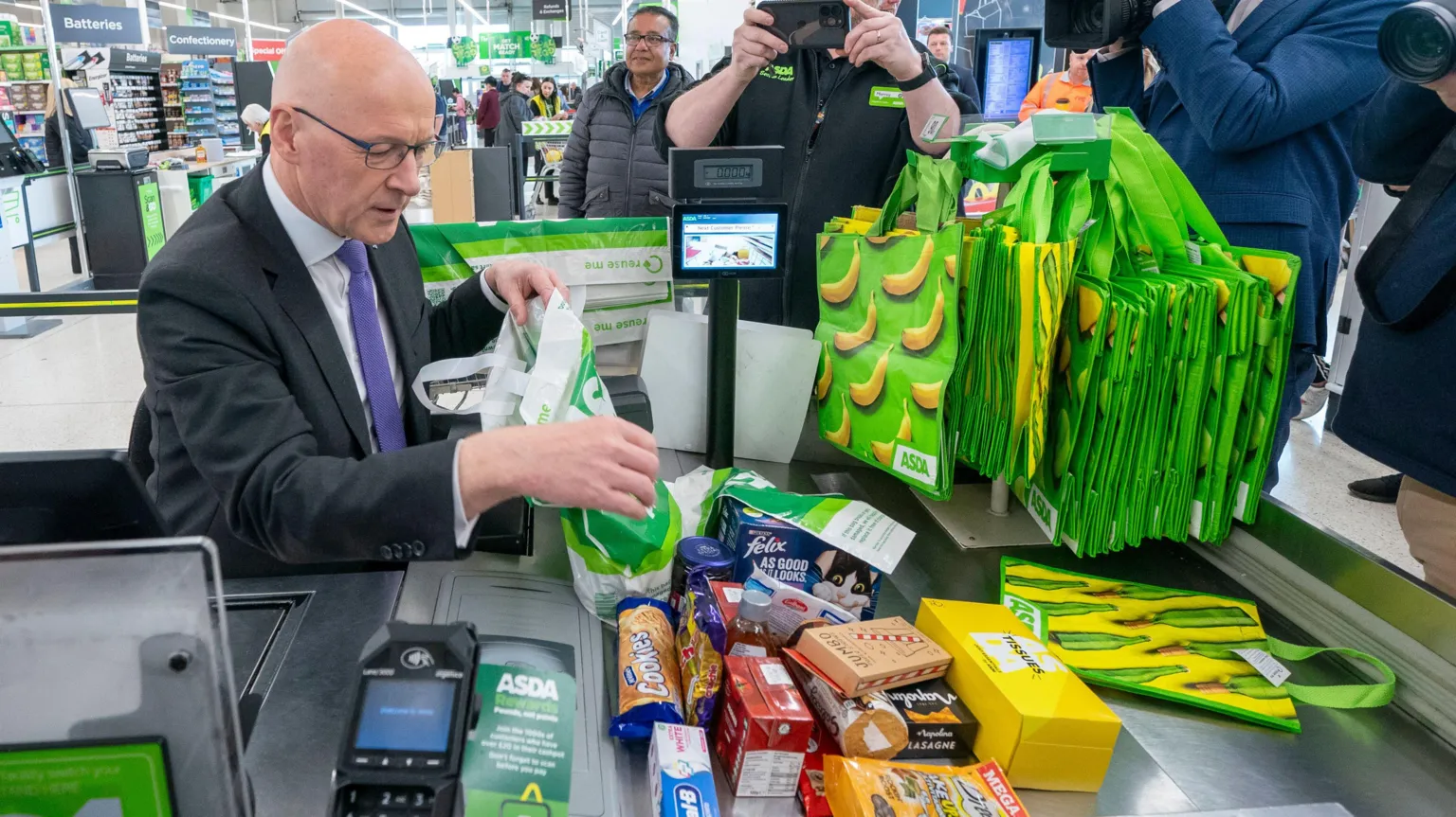 John Swinney is sitting at an Asda checkout where he is wearing a dark suit, white shirt and purple tie. He is bagging a pile of shopping as photographers and camera people capture the image.