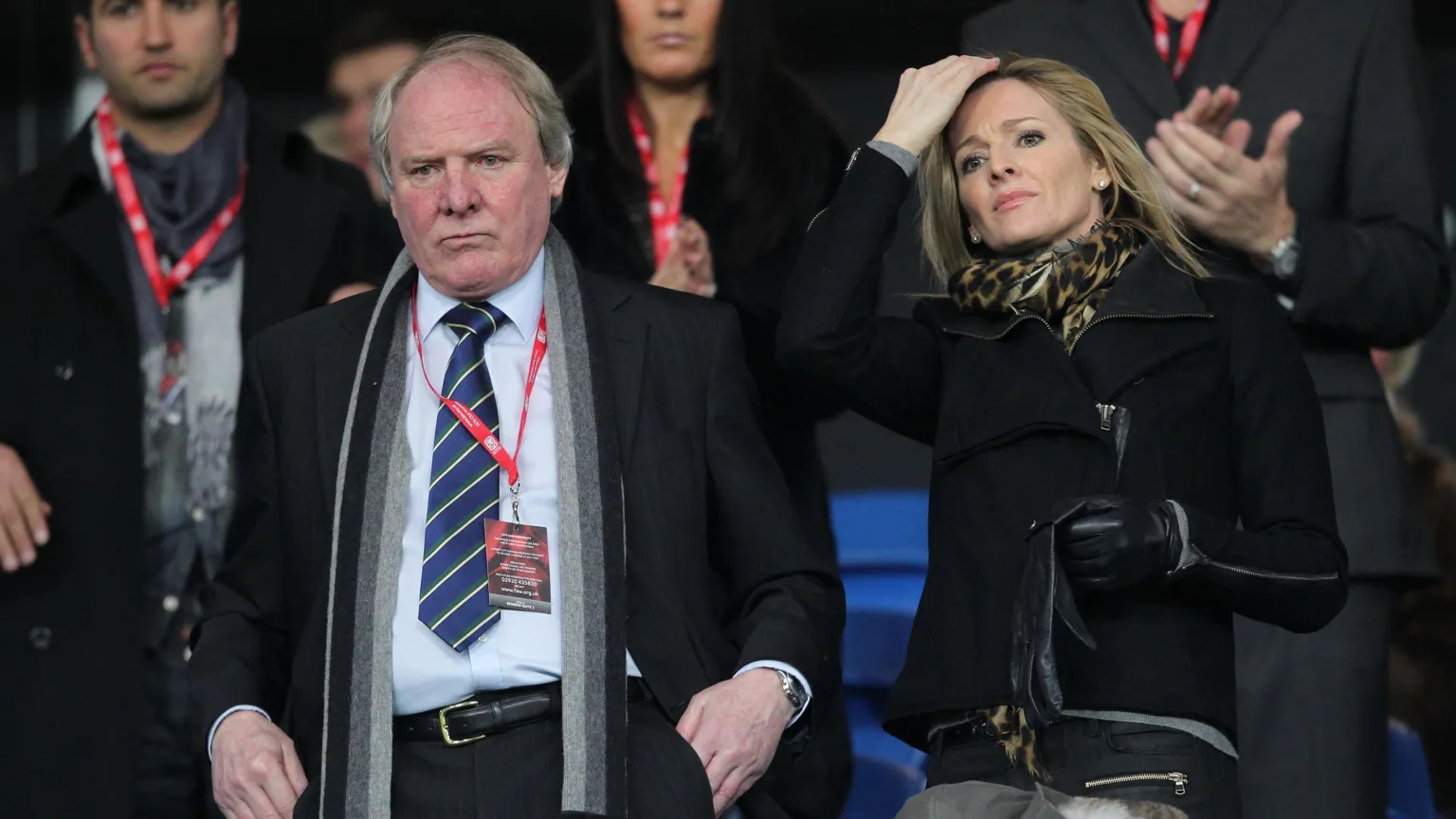 A man and woman stood side by side looking out onto a football game, with a look of concentration on both faces. The man is wearing a blue shirt and tie, whilst the woman is wearing a black coat and cheetah print scarf.