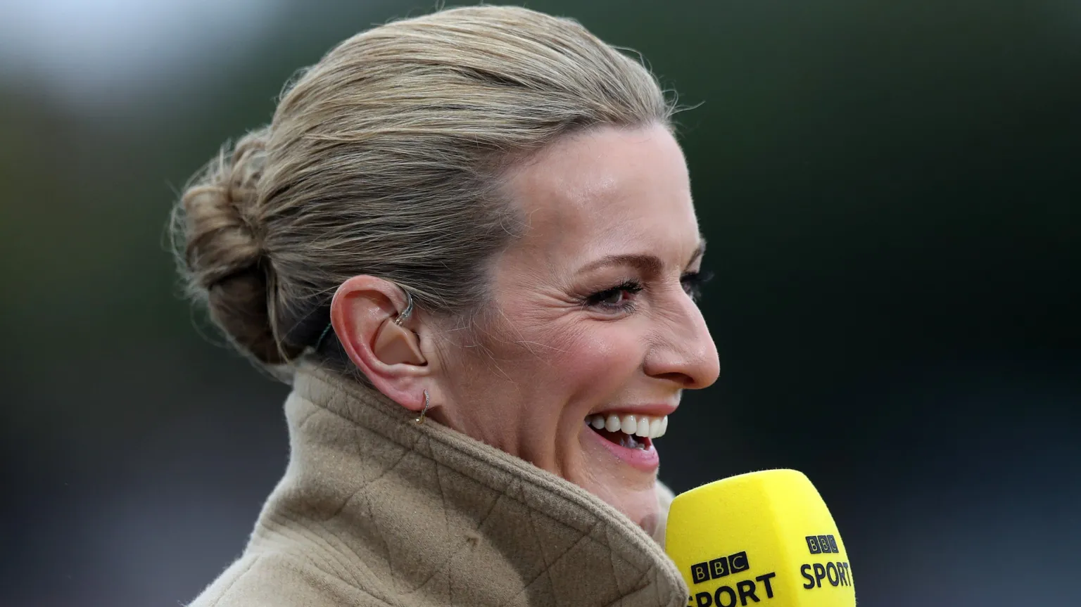 A blonde-haired woman with a happy face, wearing a beige jacket with the collar turned up. She is smiling and looking off into the distance while holding a microphone.