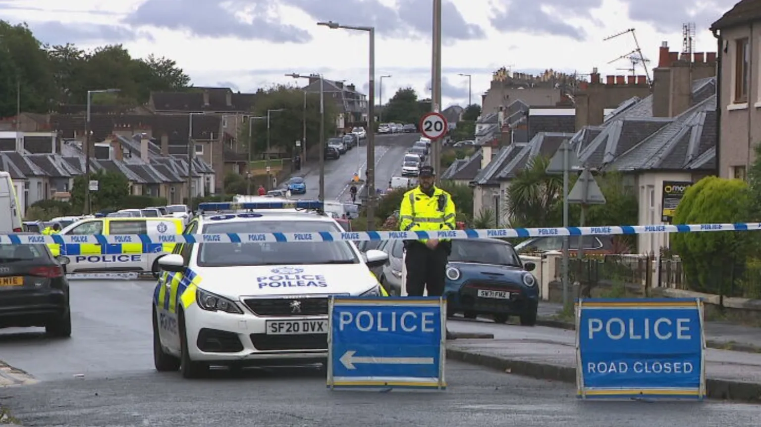 A residential street in Edinburgh cordoned off with police tape. There are blue signs that read police road closed and several police cars and vans behind the tape while one officer stands guard.