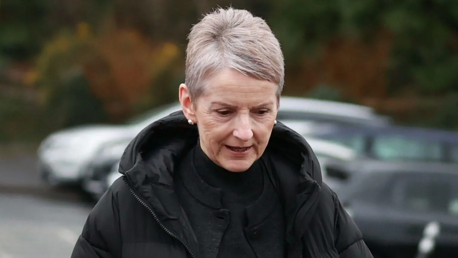 A woman with grey hair, wearing a dark top and coat. She also is wearing earrings.