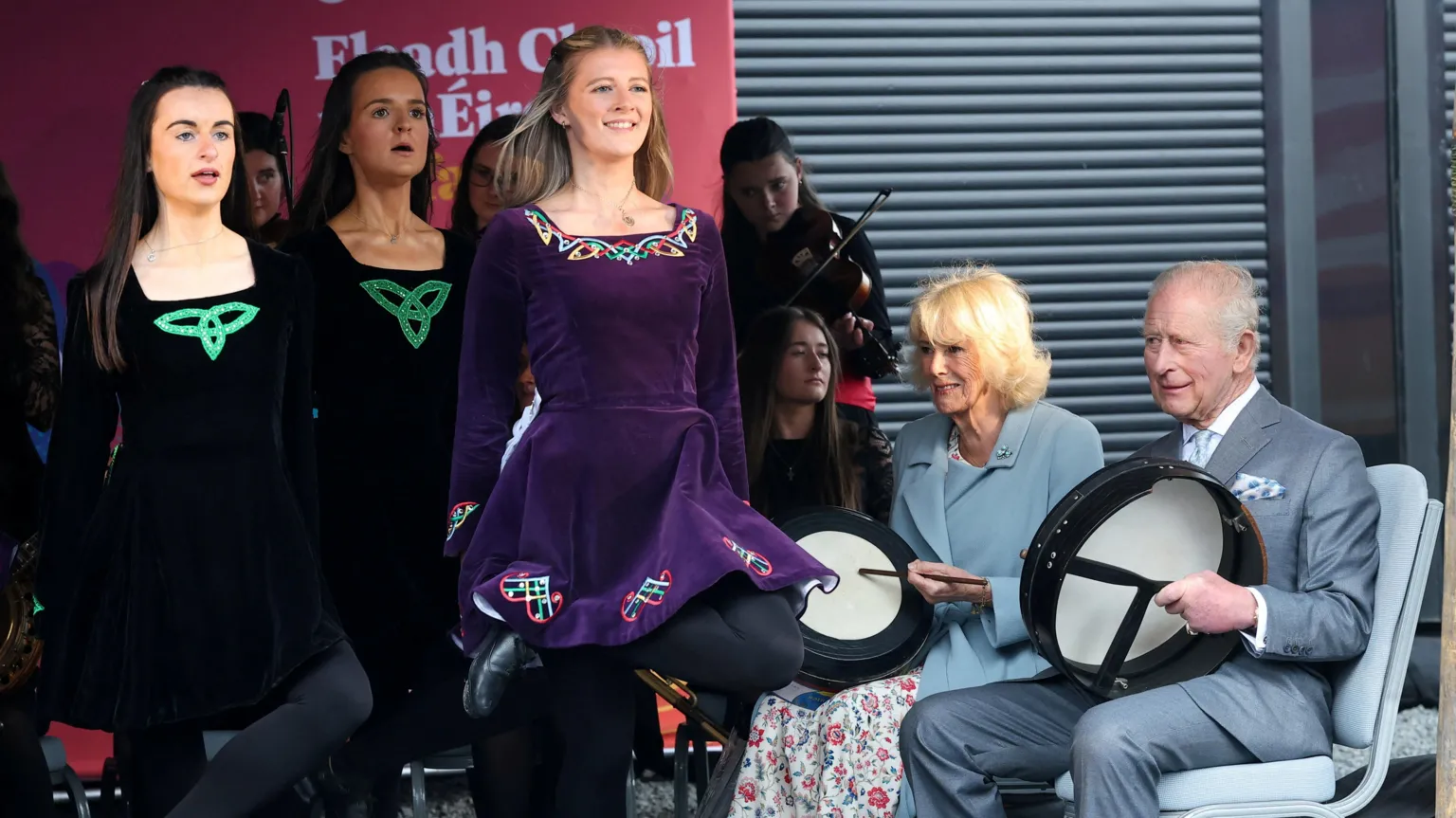 Three Irish dancers can be seen on stage. Two wearing a black Irish dancing dress and another in purple. They are seen performing for King Charles and Queen Camilla who are picture on the right of them playing bodhran drums.