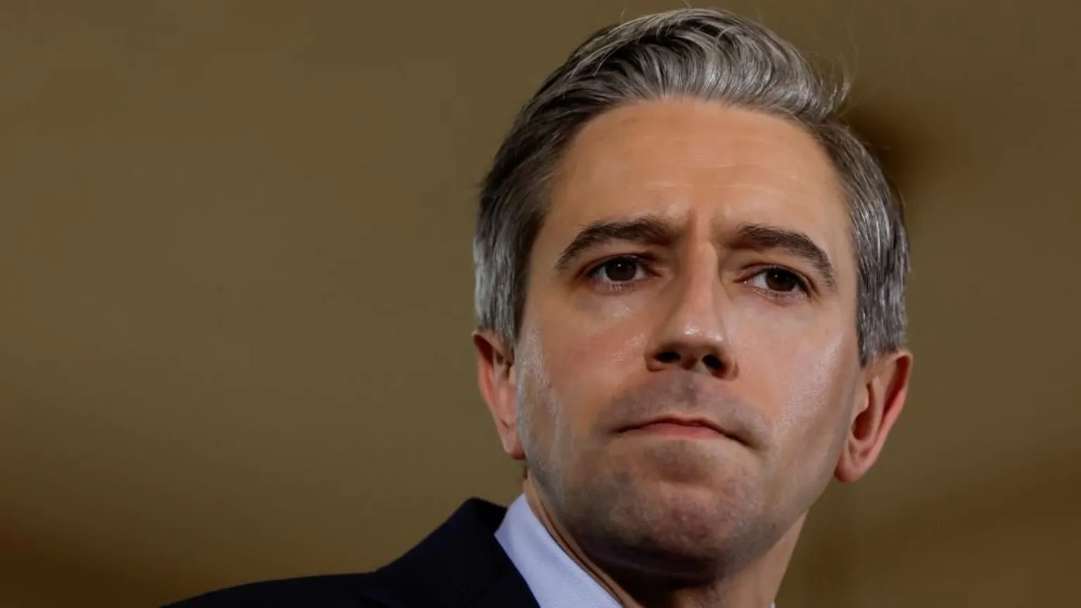  Harris is looking away from the camera. He has greying hair and is wearing a dark coloured suit jacket and a white shirt.