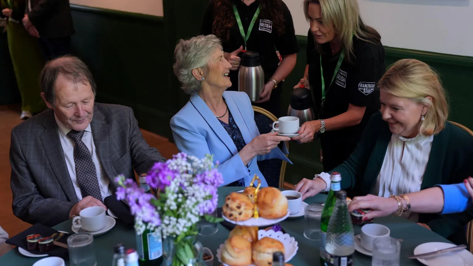 A woman with short grey hair, wearing a blue suit and navy top turns to her left and laugh's as she receives a cup of tee from a long-blonde haired woman in a dark-green polo top.