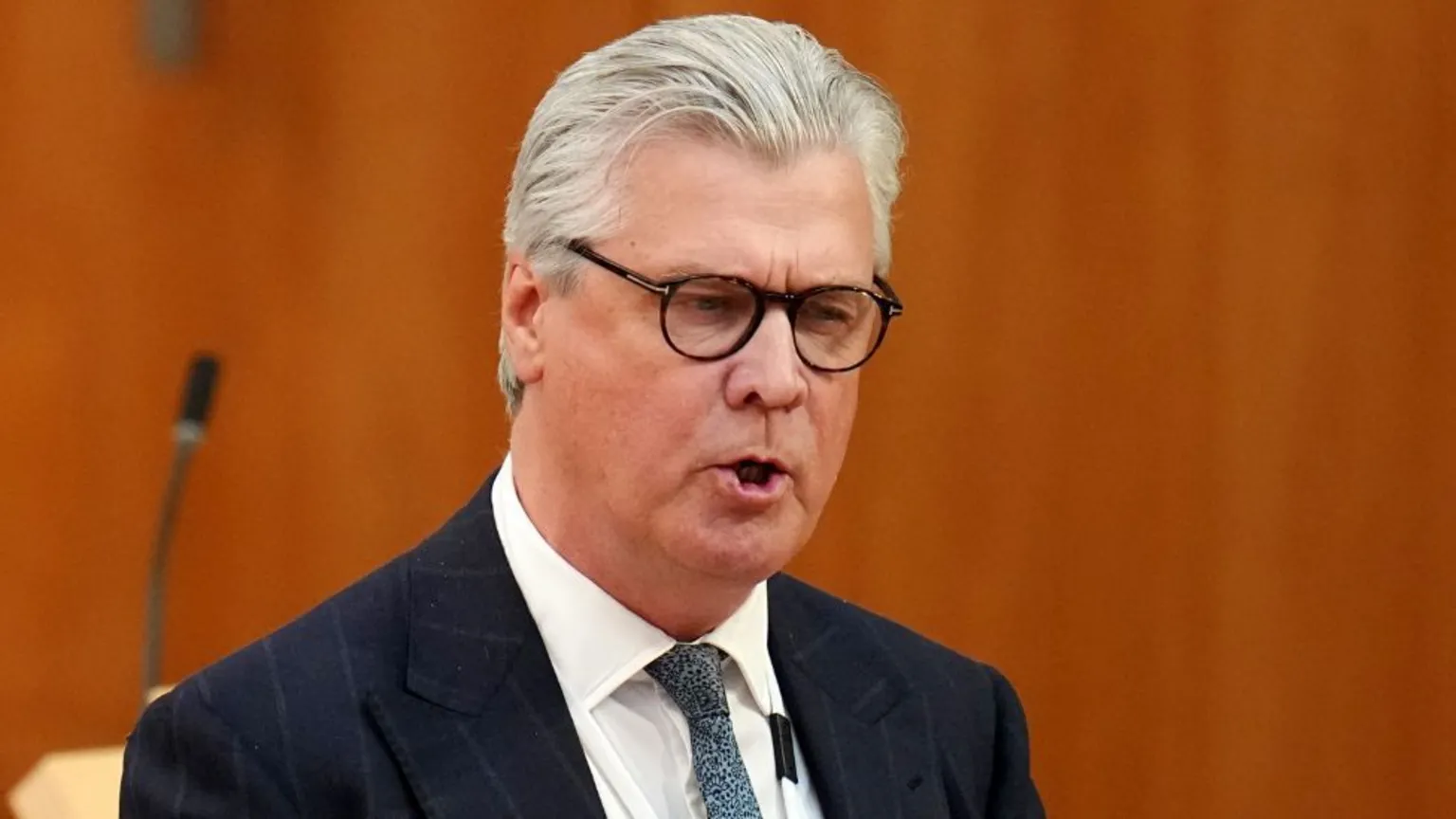 Malcolm Offord, who has grey hair swept back and black rimmed glasses. He is wearing a business suit with wood panelling behind him