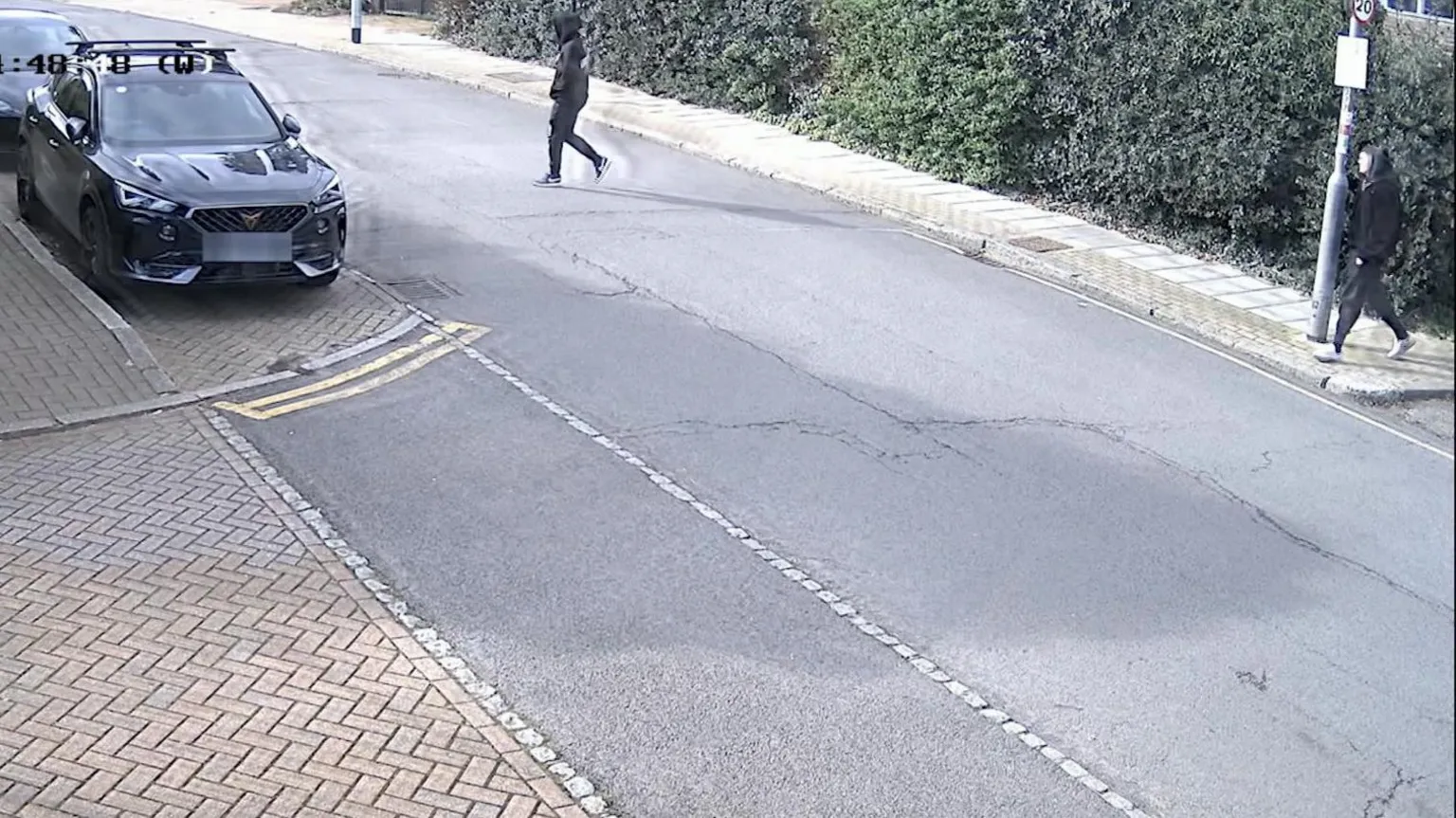 Metropolitan Police CCTV footage showing two men in black tracksuits crossing a street in Wimbledon, London. Both men have their hands in their pockets and their hoods up.