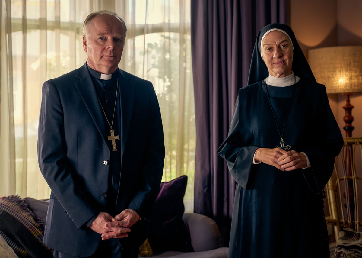 A bishop and an abbess in their robes stand in a living room.