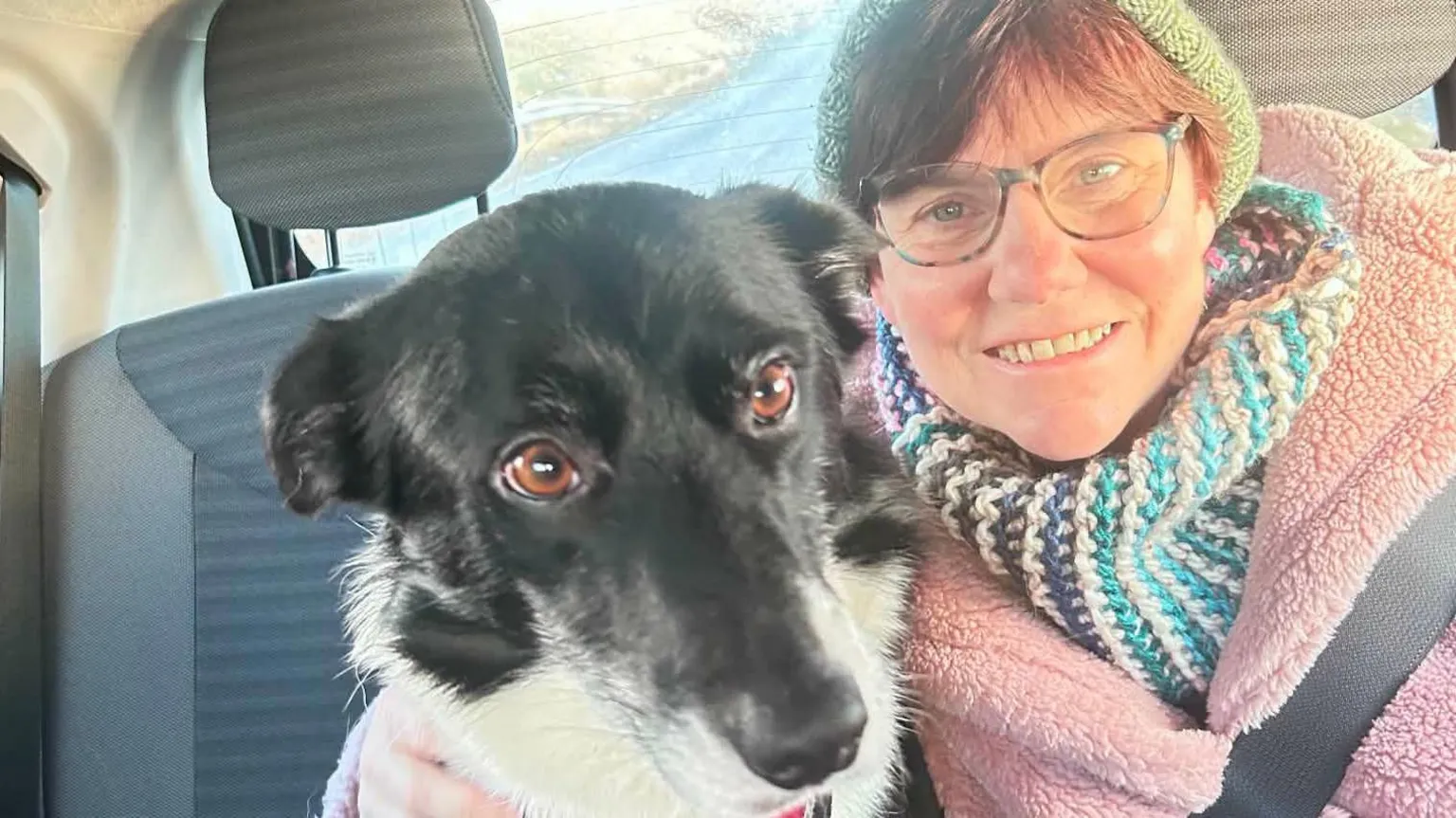 Susan Taylor A woman with red hair in a green hat with a colourful scarf and coat has her arm around a black and white dog with brown eyes. They are sitting on the front seats of a car and the woman has a seatbelt on