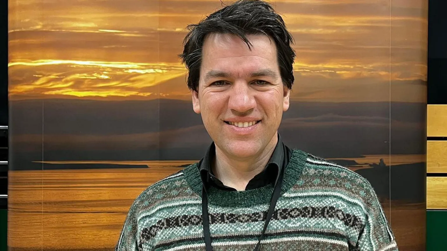 A man with dark hair and a patterned green and black jumper stands in front of a picture of a sunset