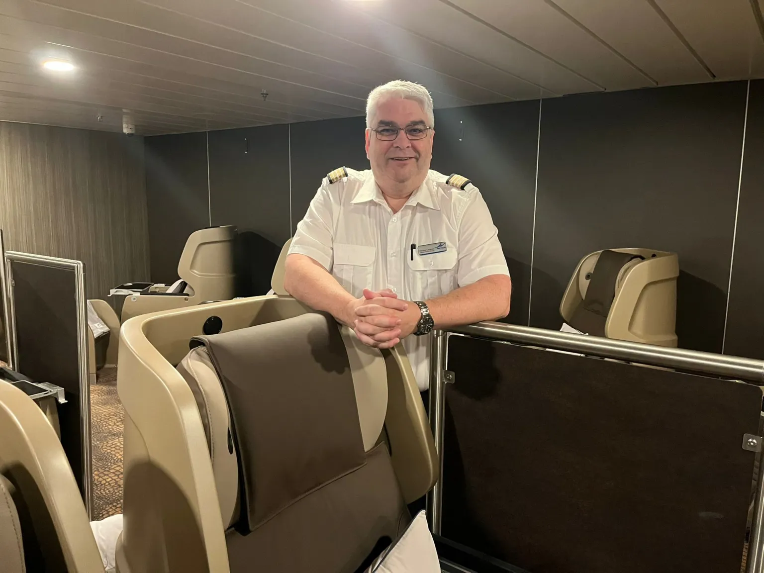 A man in a white shirt with and grey hair and glasses stands behind one of the sleeping pods which has its chair in the upright position
