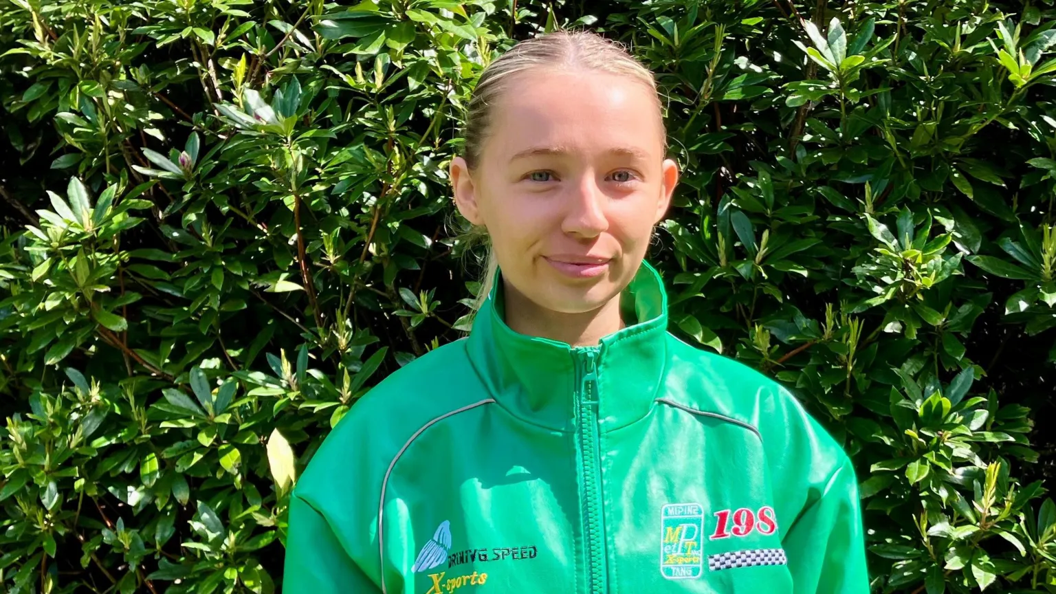 Amie Donaghy has blonde hair tied back in a ponytail and is wearing a light green driving-style jacket with a high collar. She is standing in front of a hedge.