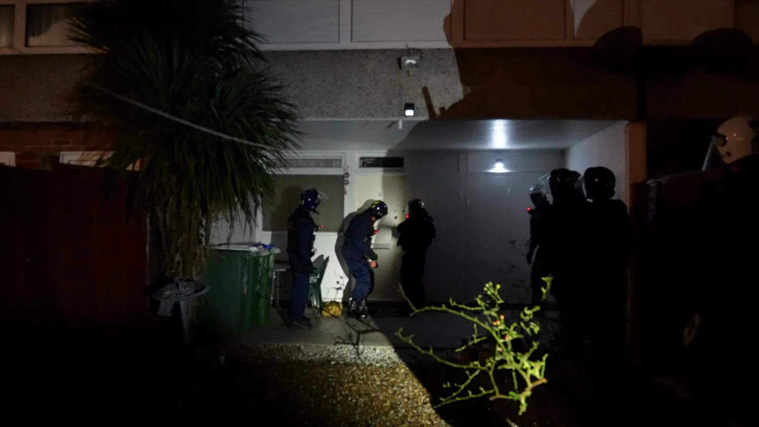 Three police officers outside a ground floor apartment at night time, dressed in helmets and riot gear, preparing to knock down the door