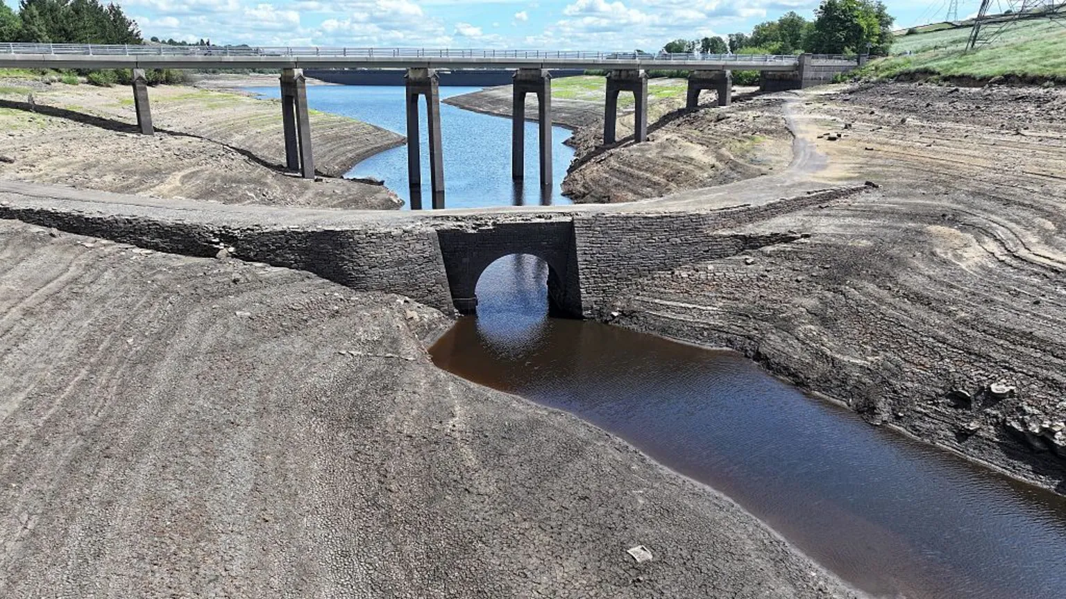 Dried-up reservoir bed, with a small stream instead of a large expanse of water. A bridge is in the background and there is a light blue sky with scattered clouds.