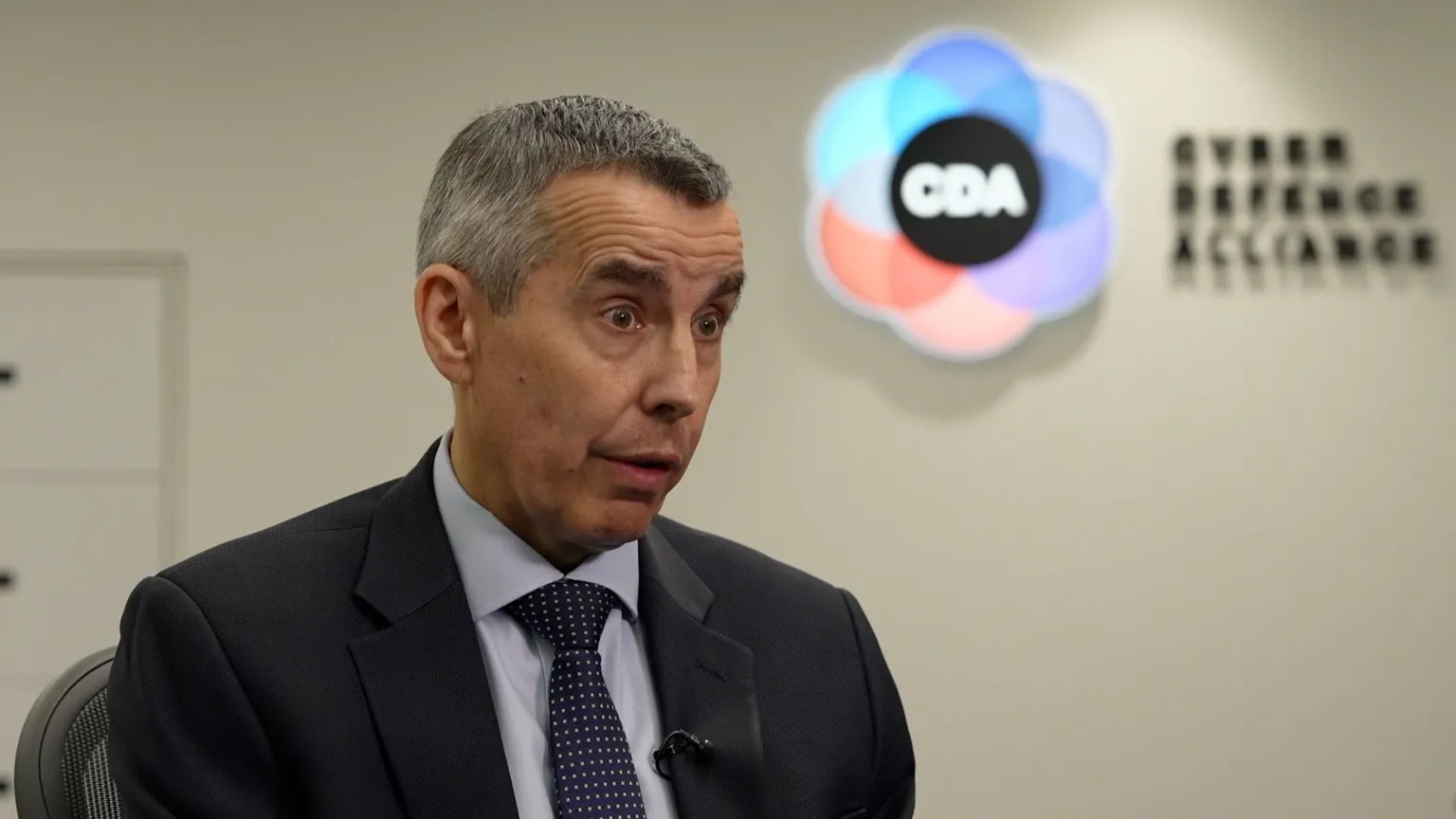The head and shoulders of a man with short grey hair, wearing a blue jacket, shirt and tie, with a CDA logo blurred in the background.