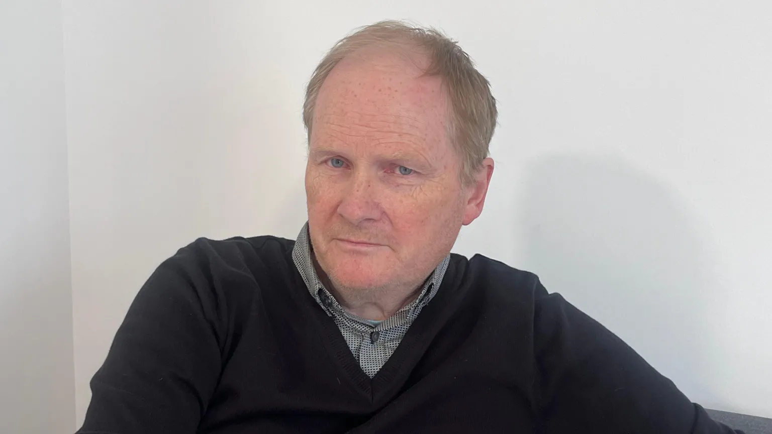 William Duncan is wearing a navy v neck jumper with a shirt underneath. He's sitting on a coach. the wall behind him is off white.