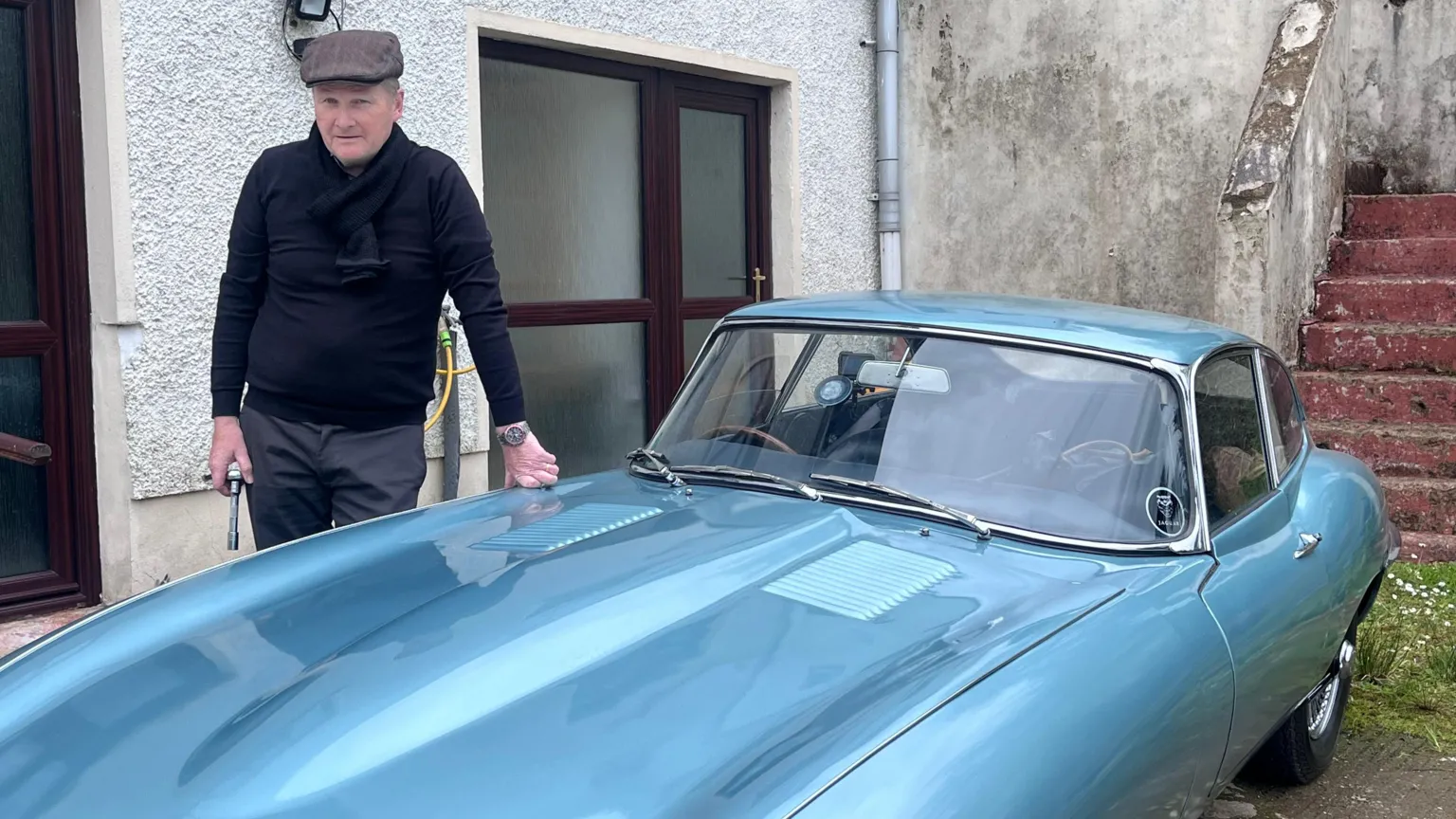 William Duncan is standing next to a blue vintage car. He is wearing a flat cap and has a navy scarf and jumper on with grey trousers. One of his hands is resting on the car and the other is holding a walking stick.