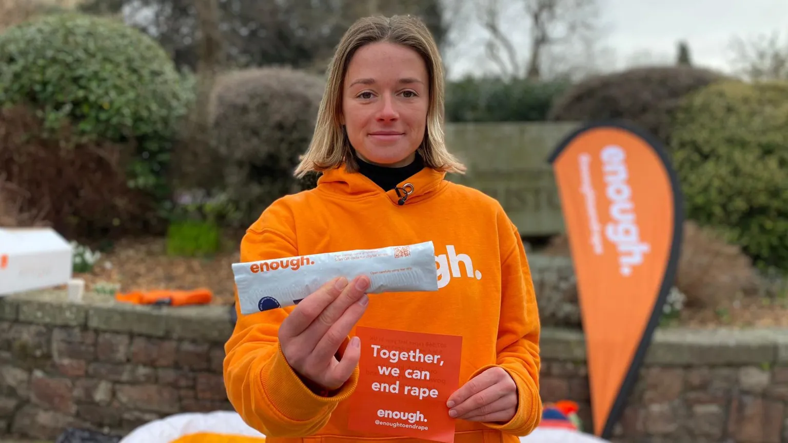 Katie White stands outside, looking to camera and holding up a self-swab DNA kit, which is inside white packaging with the word Enough on it. She is also holding a flyer that says 'Together we can end rape'. She is wearing a bright orange hoody.