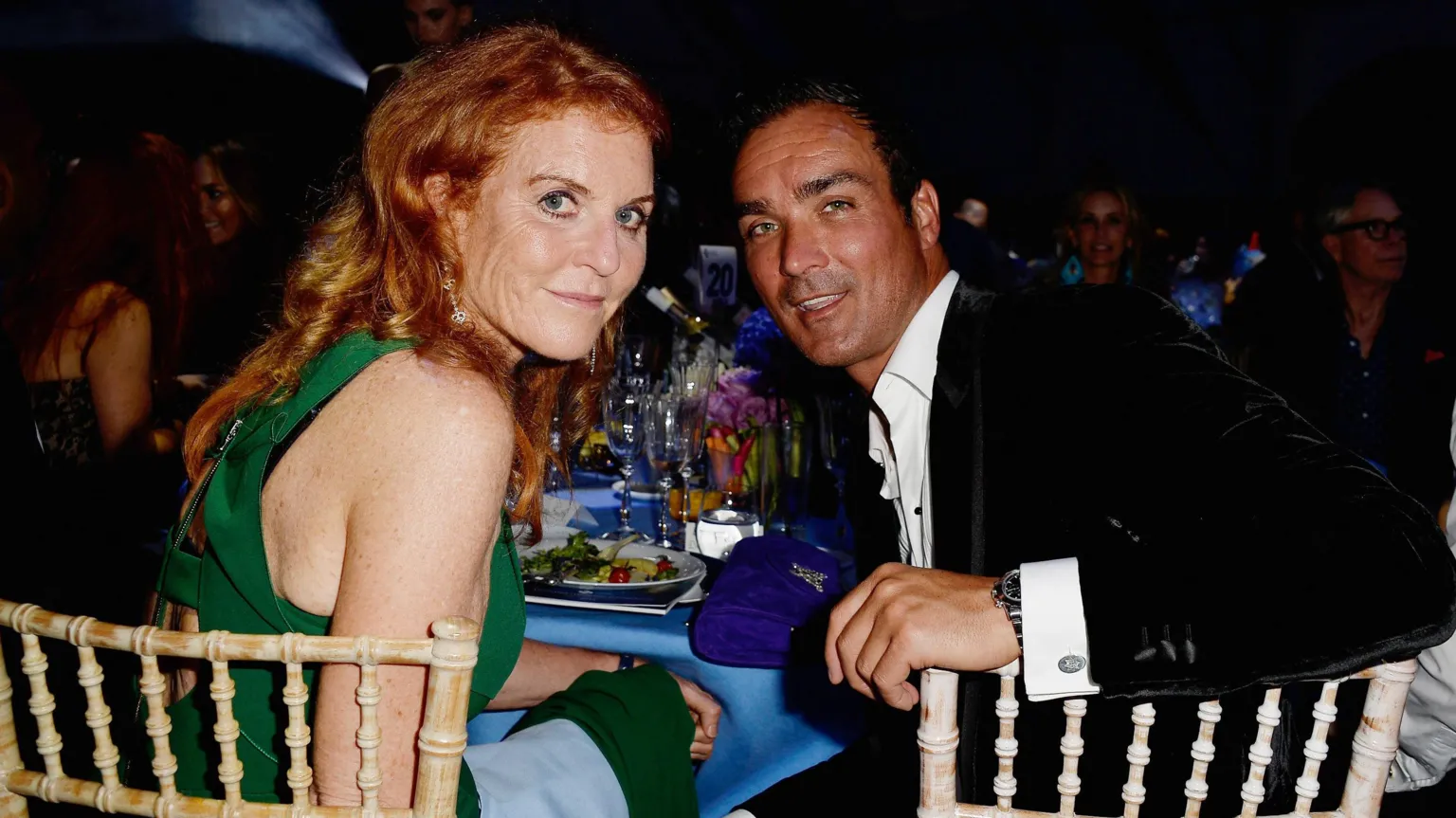 Sarah Ferguson and Manuel Fernandez sit at a table laden with glasses, plates and a blue table cloth. She wears a green dress, he wears a black velvet jacket and a white shirt open at the neck. They sit at wickerwork-style chairs, and are looking over their shoulders at the camera