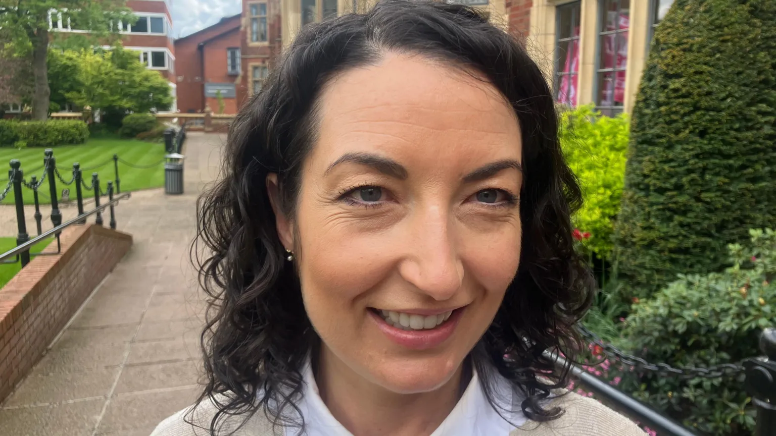 Jane smiling at the camera. She has shoulder length wavy black hair and is wearing a white shirt under a cream cardigan.
