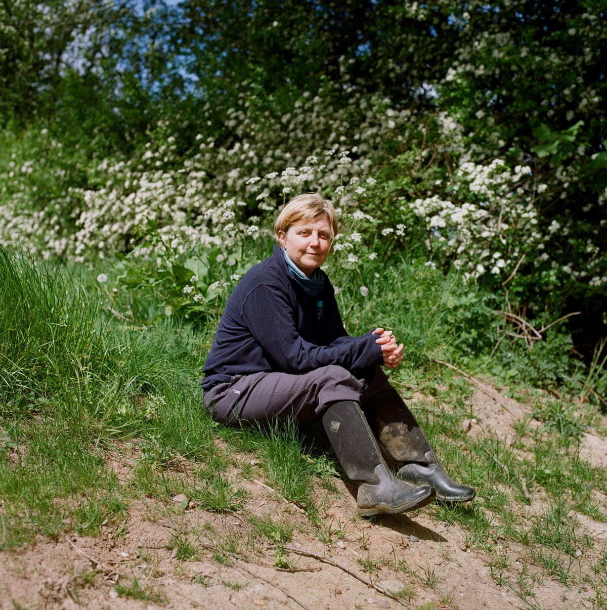 A woman with short blonde hair sits on a riverbank wearing a dark outfit with wellies