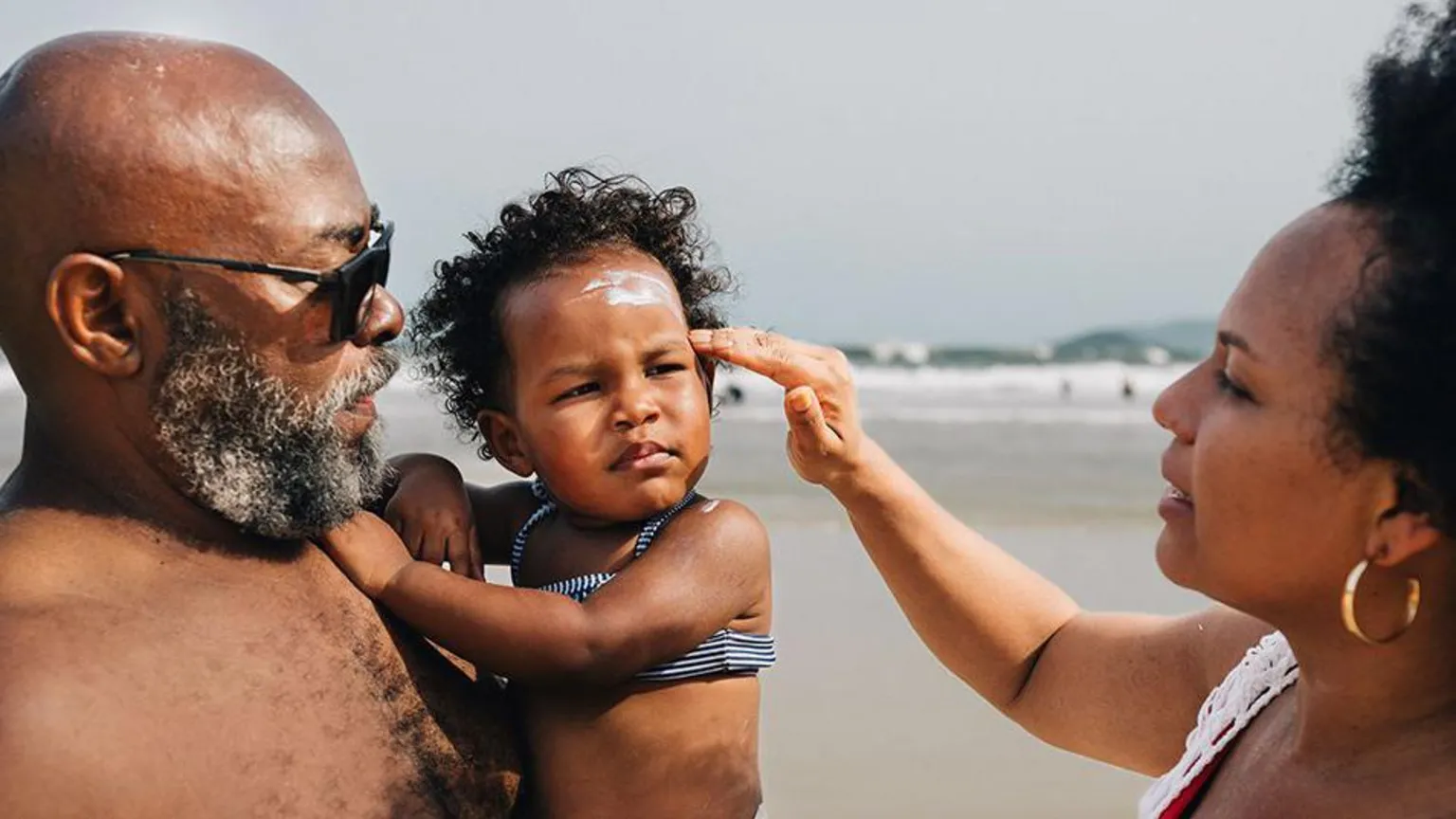  On a beach, a mum applies sunscreen to her young daughter who is in her father's arms. 