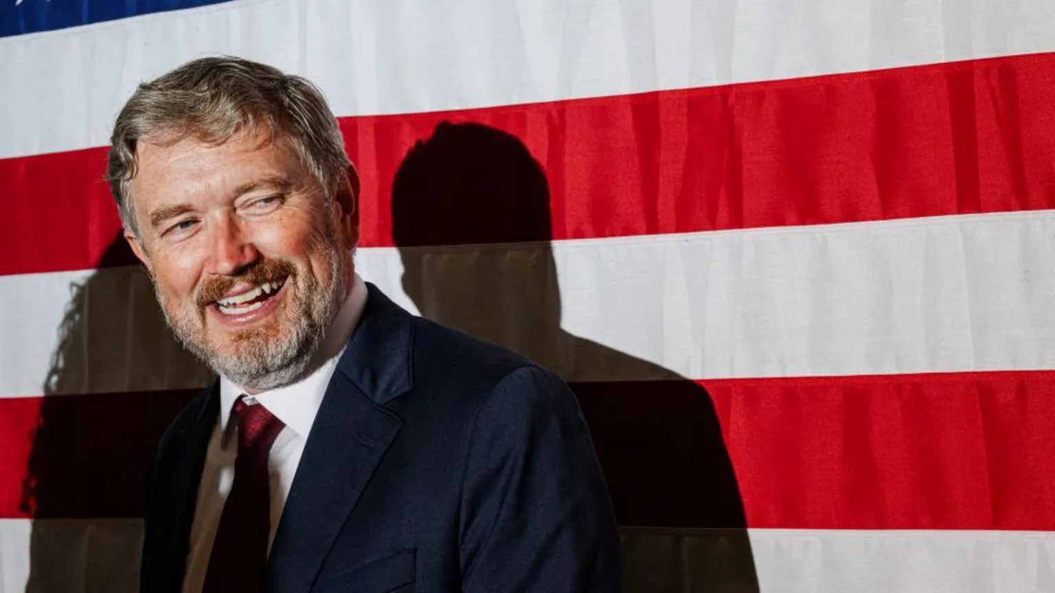 Thomas Massie dressed in dark blue suit, red tie, with a grin on his face looking to the right. He's stood in front of a large USA flag