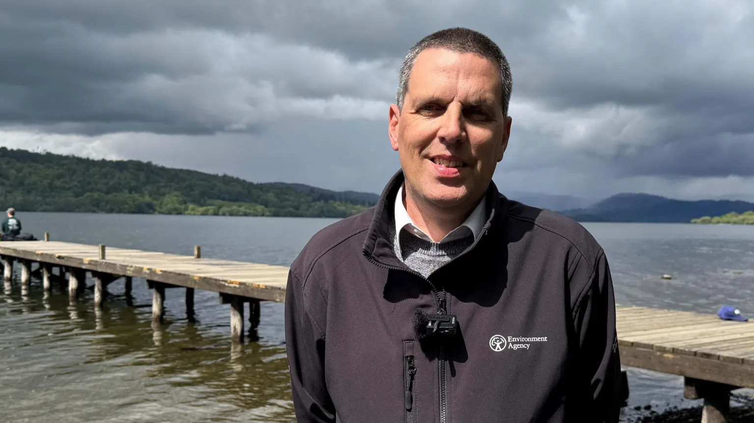 Andy Brown, water regulation manager at the Environment Agency. He has shaved grey hair and is standing in front of a small pier on Windermere. He is wearing a blue fleece with the Environment Agency logo, over a grey jumper and white shirt. He is smiling at the camera.