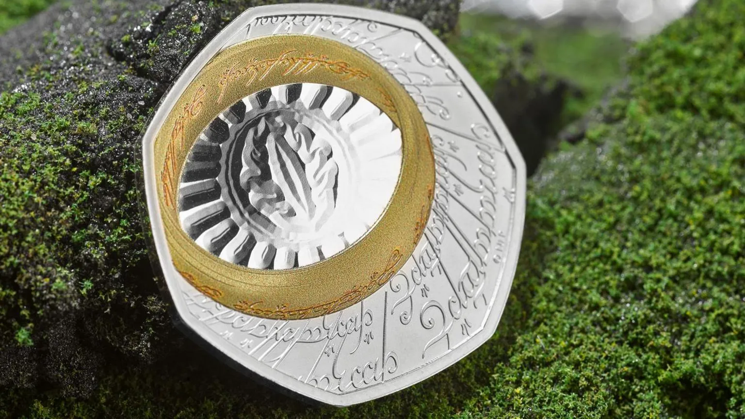 The Royal Mint A single coin on a fake background of granite grey rock frosted in green lichen. the coin is silver with an etched inscription in Elfish, a Lord of the Rings script and language. A gold band, the one ring, also inscribed in Elvish, is at the top left of the seven sided coin. the ring surrounds the eye of sauron etched onto the silver coin with an oval around the eye which is surrounded by a fan of lines to the ring. 
