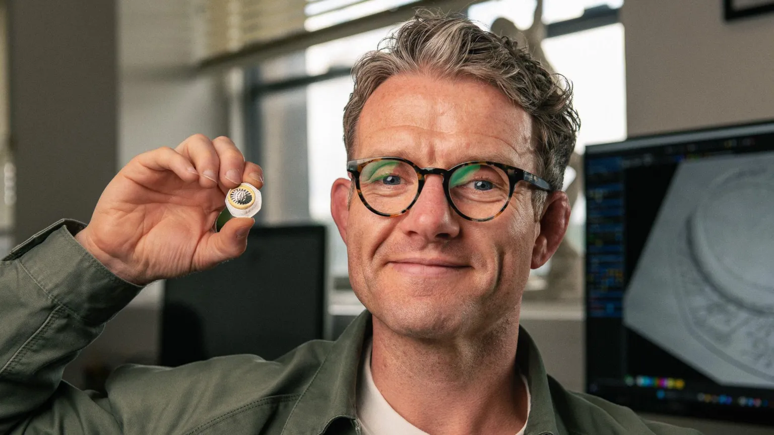 The Royal Mint A man in a green shirt holds the coin in a one hand next to his face with a graphic design of the coin in black and white on a screen behind him. he has brown and grey hair, swept back, tortoise shell rimmed glassed and is smiling for the camera 